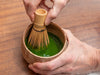 Person using a bamboo whisk to stir green tea in a ceramic bowl on a wooden surface. Koicha