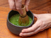 Person whisking matcha green tea in a black bowl on a wooden surface