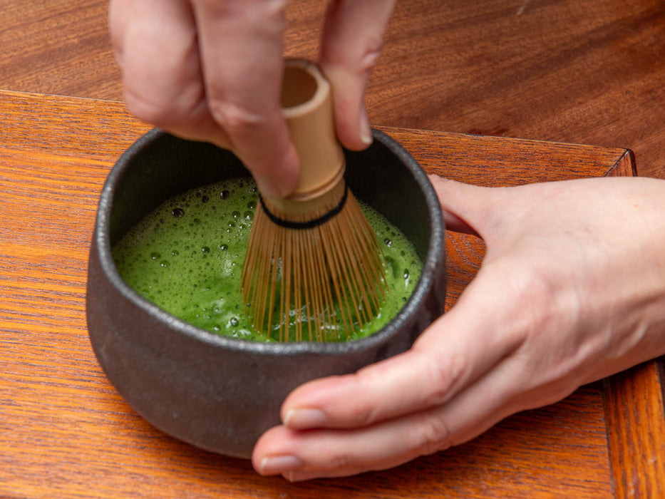 Person whisking matcha green tea in a black bowl on a wooden surface