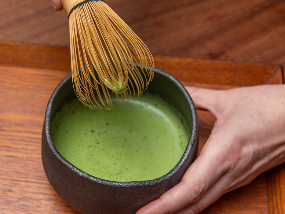 Person stirring matcha green tea in a bowl with a whisk on a wooden surface usucha