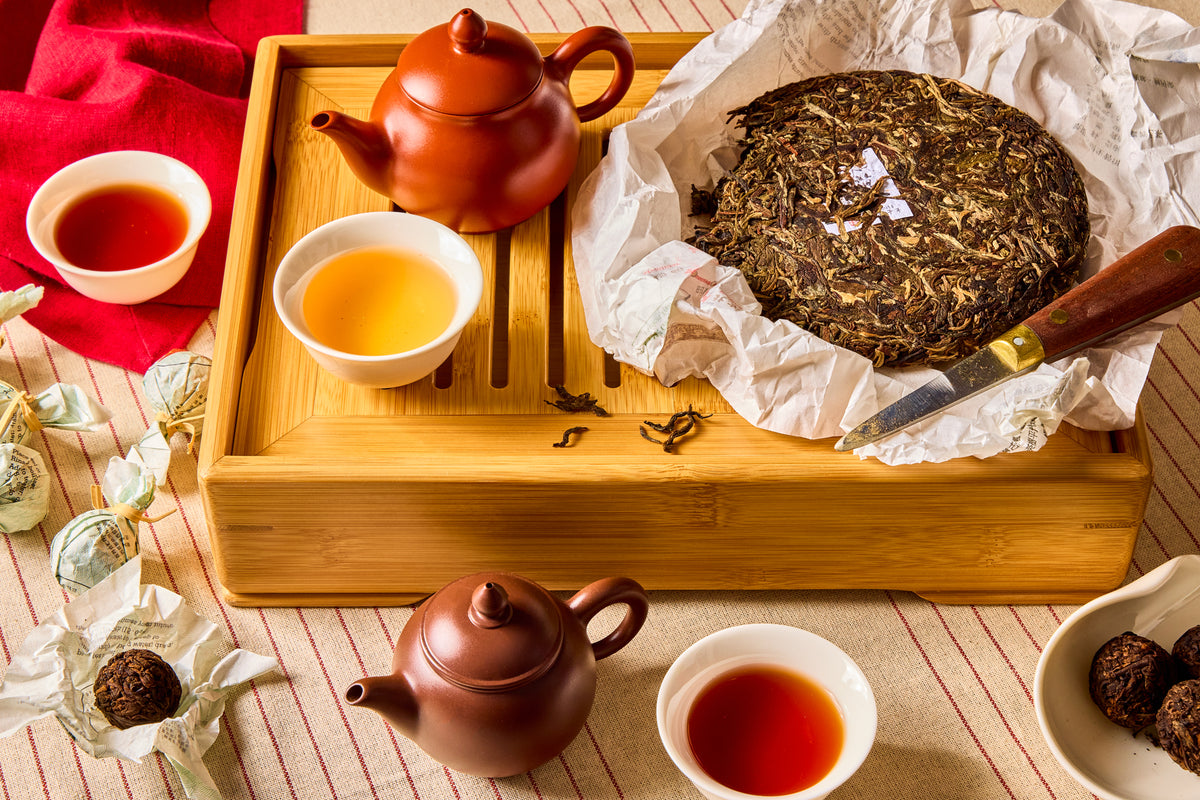 Tea-making setup with teapots, cups, and a large tea cake on a wooden tray.